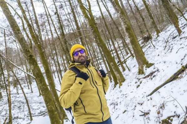 Man wearing a yellow beanie and jacket, sunglasses, and backpack, standing confident and looking at