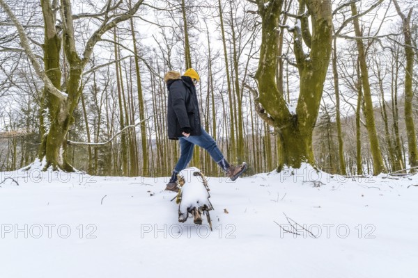 Man wearing winter clothing and a yellow beanie balancing on a snow covered log, enjoying the cold
