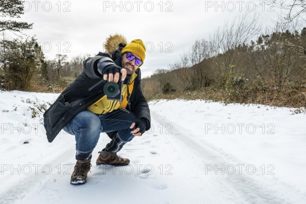 Man in winter gear with a yellow hat and sunglasses crouching on a snowy trail, aiming a camera to