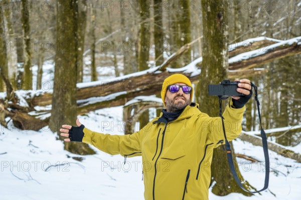 Man wearing a yellow jacket and beanie capturing moments with his camera in a snow covered forest,