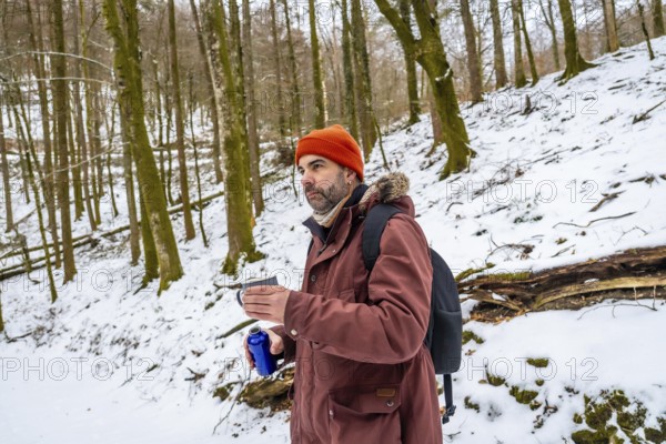 Adventurous man in a beanie and jacket pauses on a snowy forest trail with a backpack, sipping a