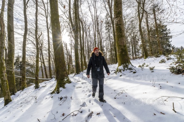 Man walking a snowy forest trail, sunlight filtering through bare trees as he hikes in warm gear,