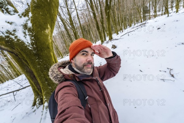 Man posing for a selfie in a snowy forest during winter, looking out with his hand shading his