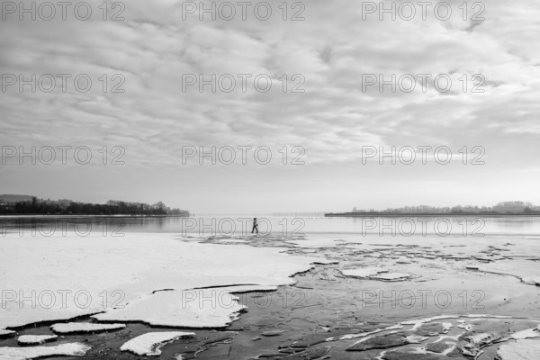 A person walks on a frozen lake under a cloudy sky in a wintry landscape, black and white