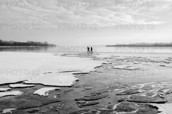 Two people walking on a frozen lake under a cloudy sky in a wintry landscape, black and white
