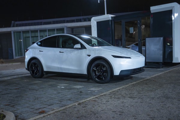 White Tesla parked at night at a charging station on grey asphalt, New Model Y Standard, Tesla