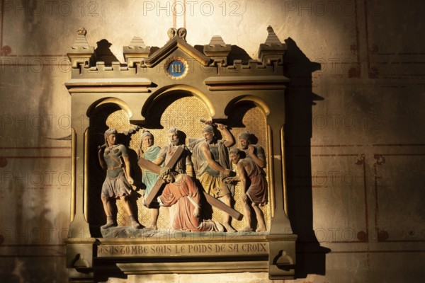 A carved relief depicts a religious scene. Jesus falls while carrying the cross. Roman soldiers and