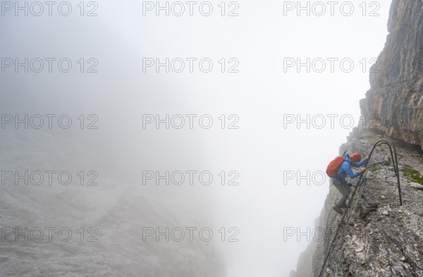 Mountaineer climbs a ladder on the Via Ferrata Oliva Detassis via ferrata in fog, cloudy steep rock