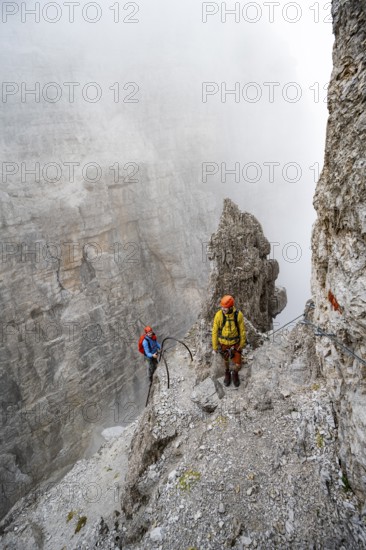 Two mountaineers climb the Via Ferrata Oliva Detassis via ferrata in fog, steep mountains covered