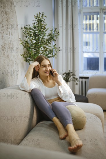 Barefoot woman is seated on a couch in a room with soft lighting. She is smiling while talking on