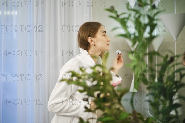 Cute woman in white bathrobe stands in a bathroom with cotton sponge in hand surrounded by green