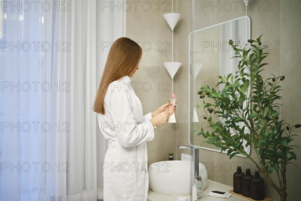 A woman stands in a bathroom wearing a robe while reading the details on a skincare product. She is