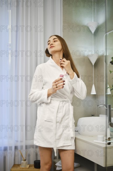 Woman stands in a bathroom wearing a robe. She holds a bottle of fragrance and is applying it to