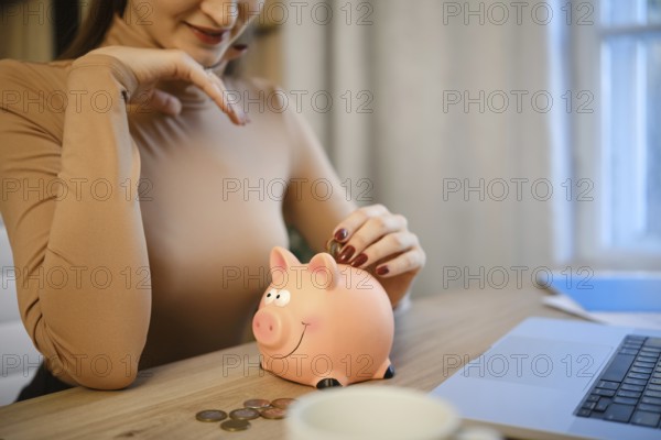 A woman sits at a wooden desk, placing coins into a pink piggy bank. She is focused on saving money