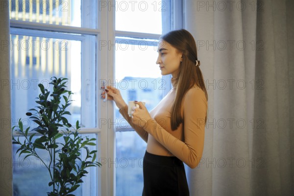 Cute woman stands by a window in a city apartment holding a coffee cup. She looks outside as the