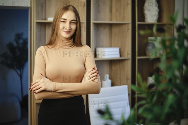 A woman with long hair stands in a modern room with wooden shelves. She has her arms crossed and