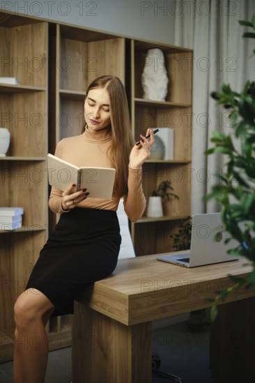 A woman sits on a wooden desk in an office. She is writing in a notebook while holding a pen. A