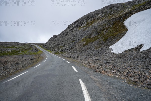 Lonely road snakes through rocky mountain landscape with snow under cloudy sky, Båtsfjord,