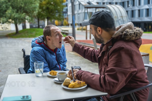 Man tenderly feeding a young boy in a wheelchair at an outdoor cafe, representing care, support,