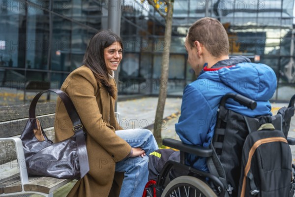 Woman sitting on bench chatting and laughing with a boy in a wheelchair in a city park, warm