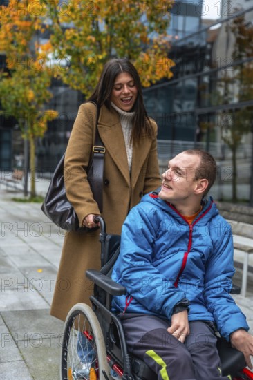Cheerful caregiver standing behind a young man in a wheelchair, pushing him along an urban sidewalk