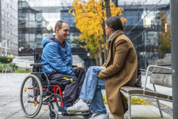 Young man living with disability in a wheelchair smiling and talking with a woman sitting on a park