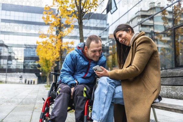 Man in a wheelchair and woman sitting on a bench, sharing a laugh and looking at a mobile phone