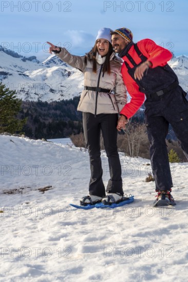 Smiling couple snowshoeing across snowy mountain slopes, pointing toward a breathtaking winter