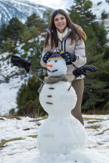 Young woman smiling while creating a snowman with a hat, scarf, carrot nose, and tree branch arms,