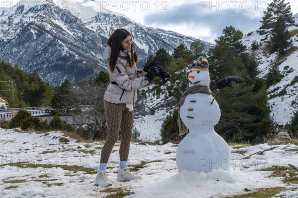 Young woman actively building a snowman outdoors with snow capped mountains and pine trees in the
