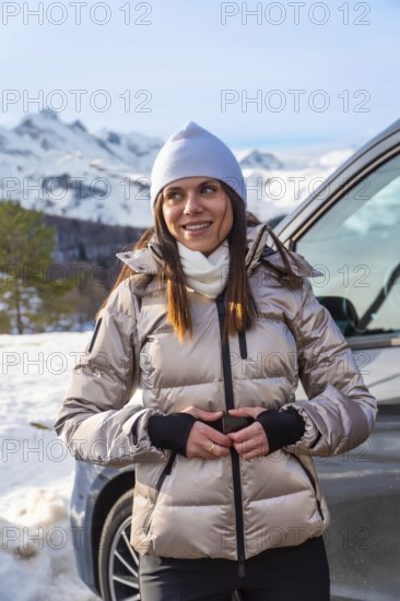 Woman wearing a puffer jacket and beanie smiling while standing by a camper van, enjoying the