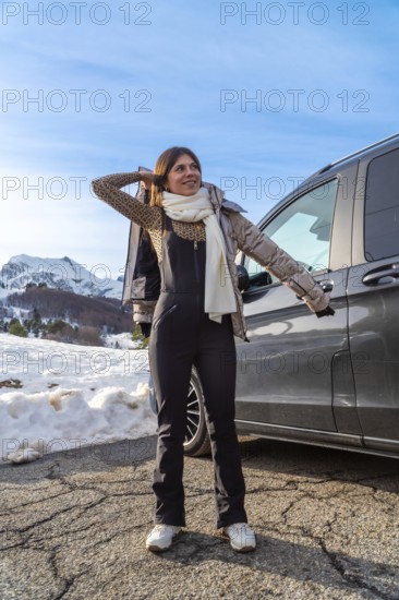 Young woman standing on a snowy mountain road next to a dark vehicle, wearing winter clothes
