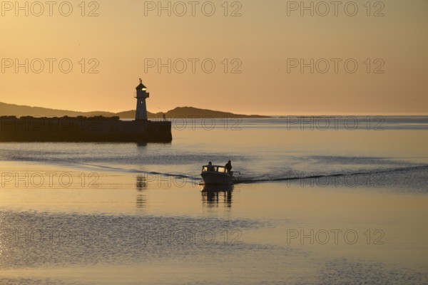 A small motorboat sails through the harbor entrance with lighthouse and midnight sun, Vardø,