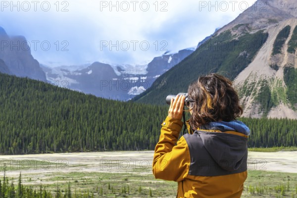 Female hiker wearing a yellow jacket using binoculars while observing a glacier in the canadian