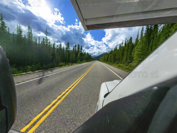 White vehicle driving along icefields parkway, surrounded by lush forests, under a bright blue sky