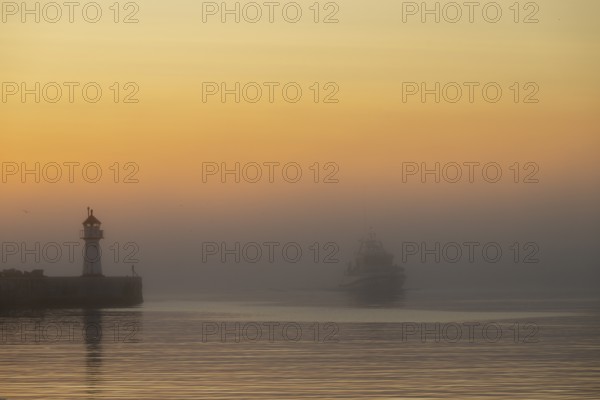 A lighthouse at the entrance to Vardö harbour at sunset with a ship in fog on calm water, Vardø,