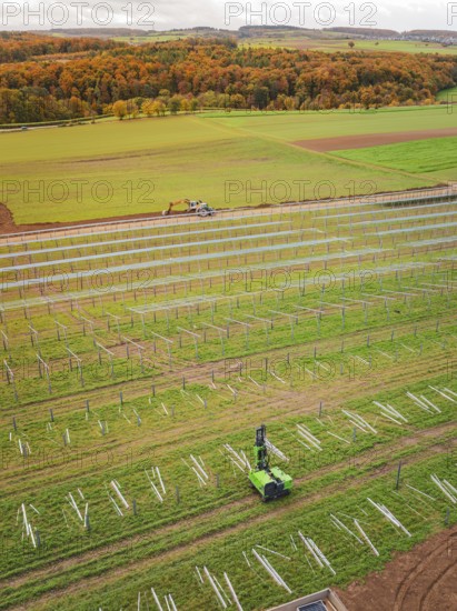 Tractor works in a green field with solar plant construction beginning, surrounded by autumnal
