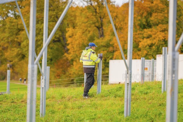 A worker wearing protective clothing erects metal structures on a grass-covered area in autumn,