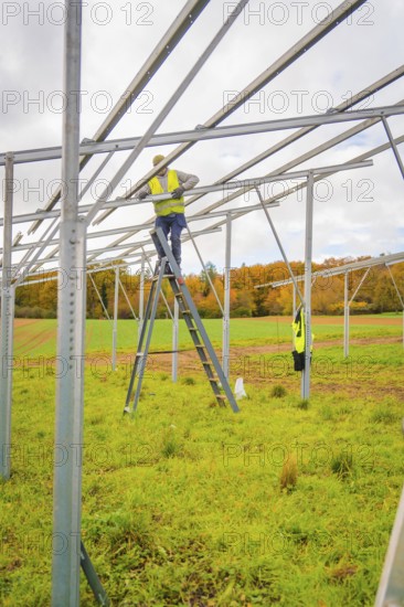 Workers stand on a ladder, assemble metal scaffolding over a green area surrounded by fields,