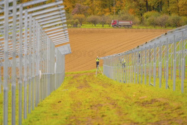 A farm worker on a ladder between metal structures with a tractor in the background in an autumn