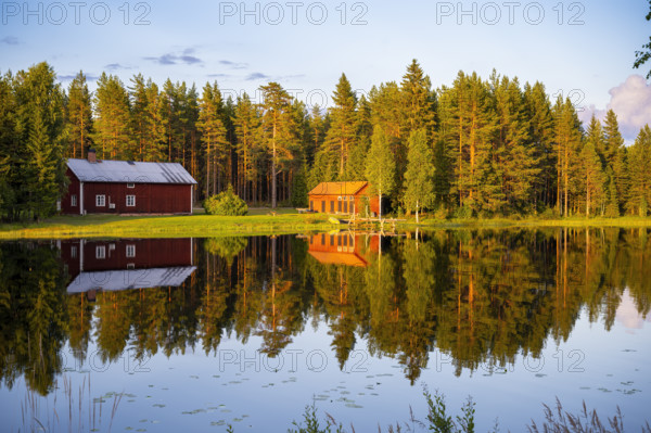 Two red houses are reflected in a quiet lake surrounded by forest under the warm light of the