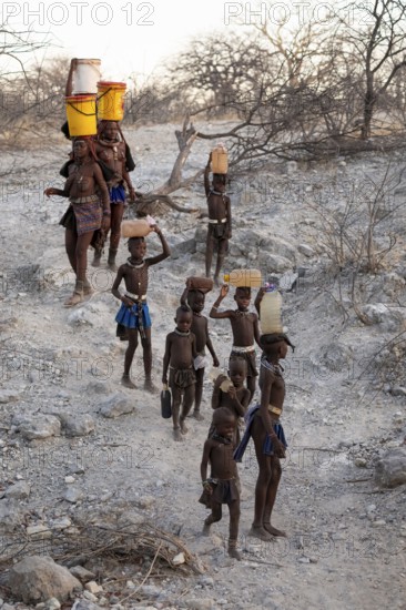 Himba woman and children run to fetch water with canisters through dry countryside, traditional