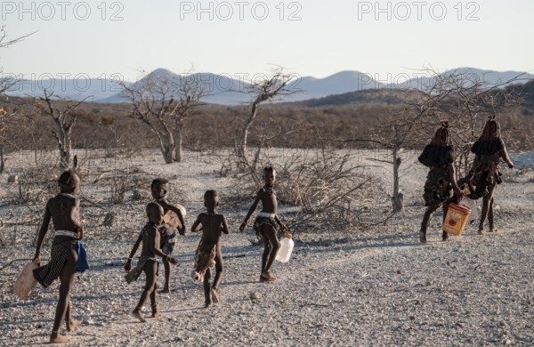 Himba woman and children run to fetch water with canisters through dry countryside, traditional
