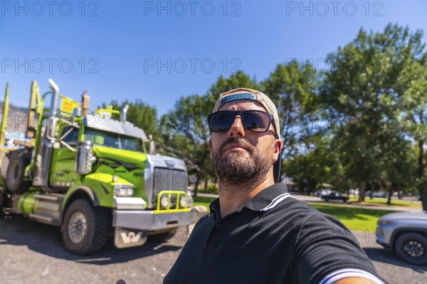 Trucker wearing sunglasses and baseball cap taking selfie with his logging truck in a parking lot