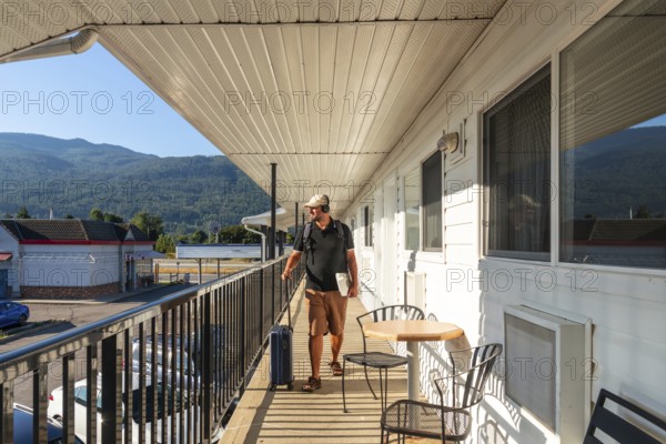 Male tourist wearing headphones and carrying a laptop strolls along a motel balcony, pulling