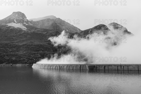 Fog rises over the dam of the Lac de Moiry reservoir, black and white photo, Val d'Anniviers,