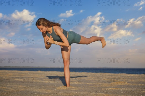 A woman performs Virabhadrasana Warrior 3 pose on the beach during sunset. Her strong stance and