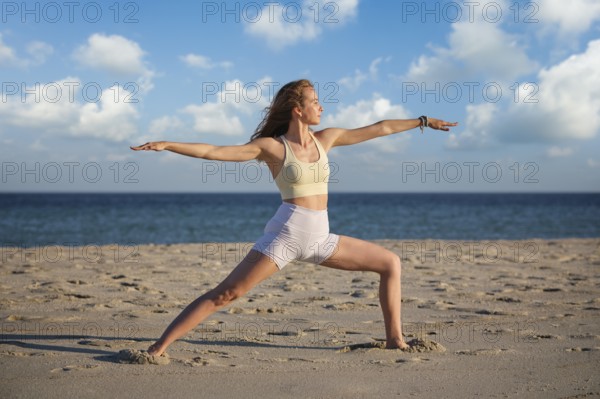 A woman performs the Virabhadrasana 2 Warrior 2 pose on a sandy beach, backlit by a beautiful