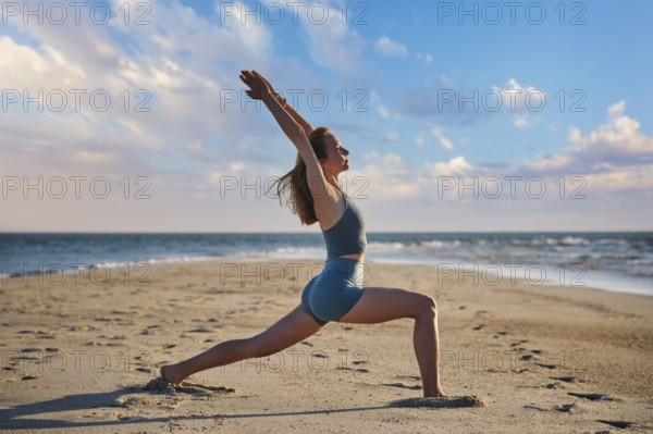 A woman performs Virabhadrasana 1, or Warrior 1 pose, on the beach during sunset. She stands strong