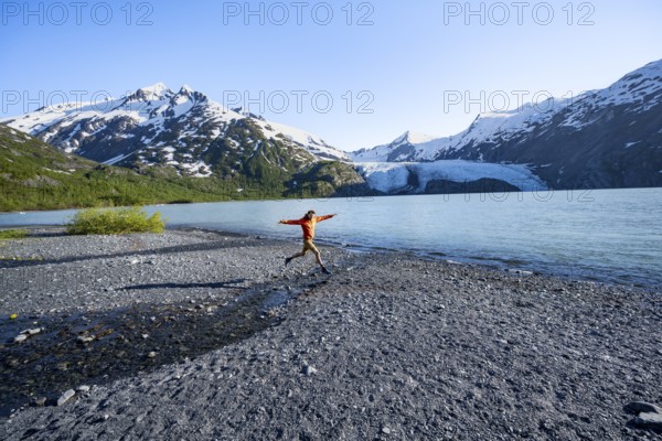 Young man jumping on a pebble beach on a glacial lake, Snowy Mountains and Portage Glacier on
